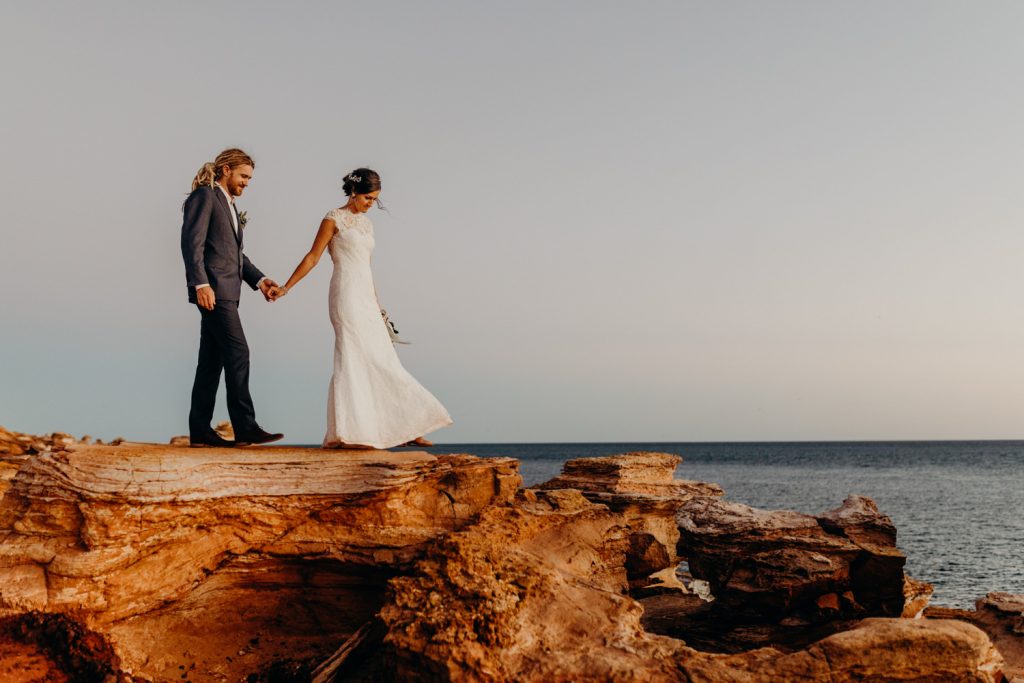 wedding couple walking on red cliff top in Broome holding hands with ocean view