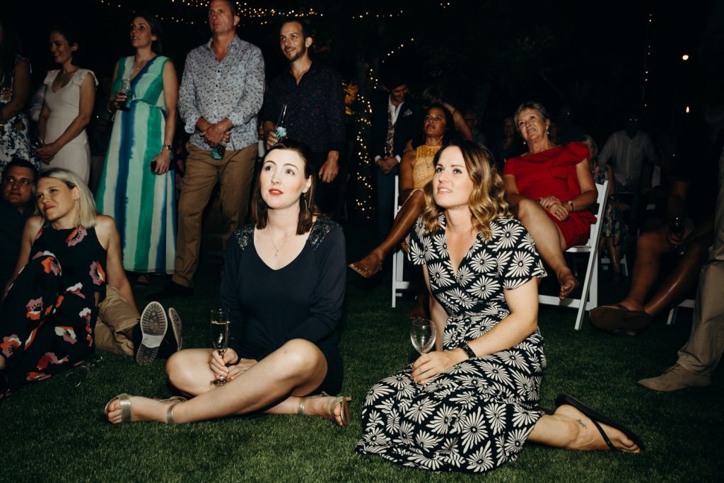two young women sitting on the lawn with other people standing around
