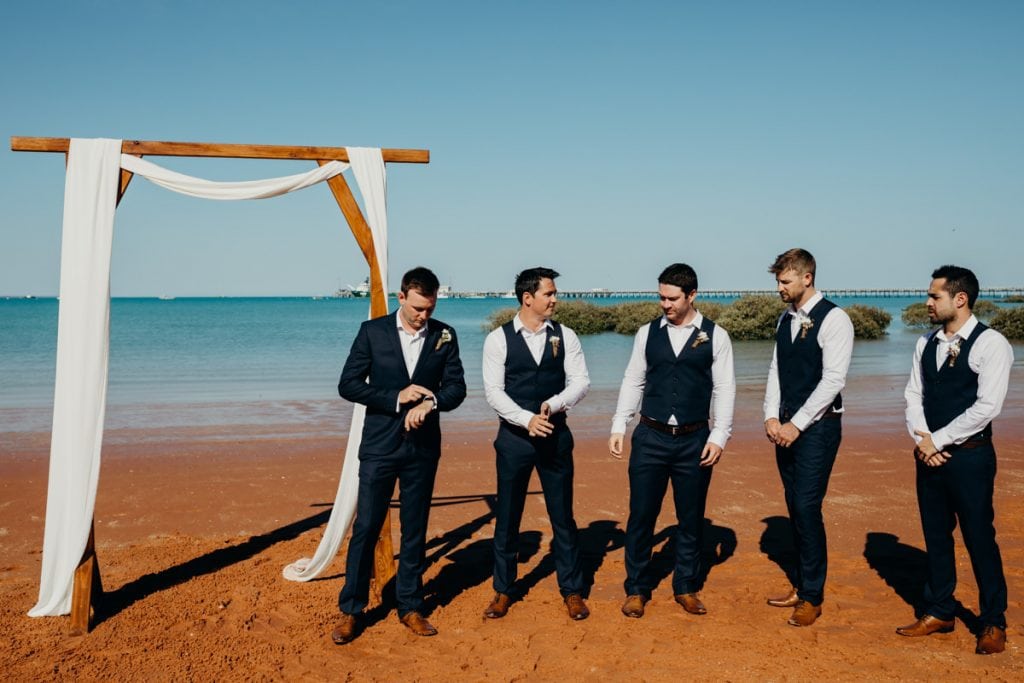 Groom and groomsmen waiting at wooden arbour by Broome Vintage Wedding Hire with Roebuck Bay in the background