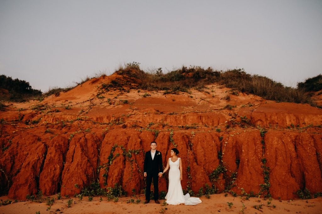 wedding couple stands in front of red cliff with bride looking at groom