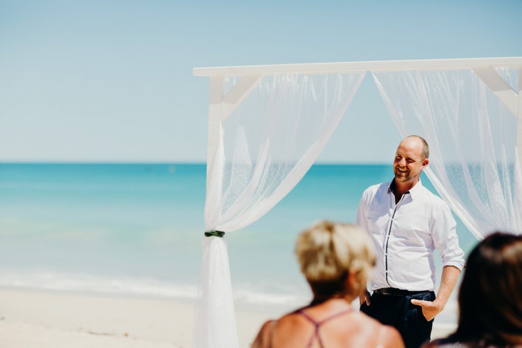groom standing under a white arbour on Cable Beach waiting for his bride