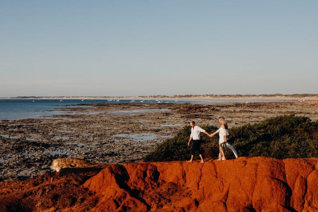 bride and groom walking along red pindan cliff with view over Cable Beach