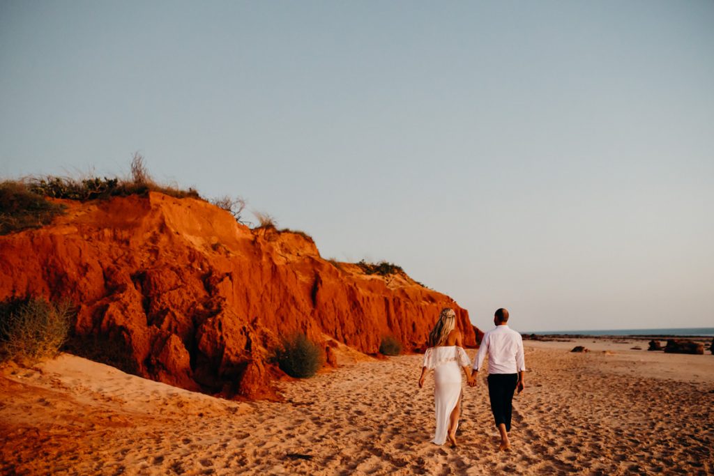 wedding couple walks hand in hand along Riddell Beach with red cliffs and Indian Ocean