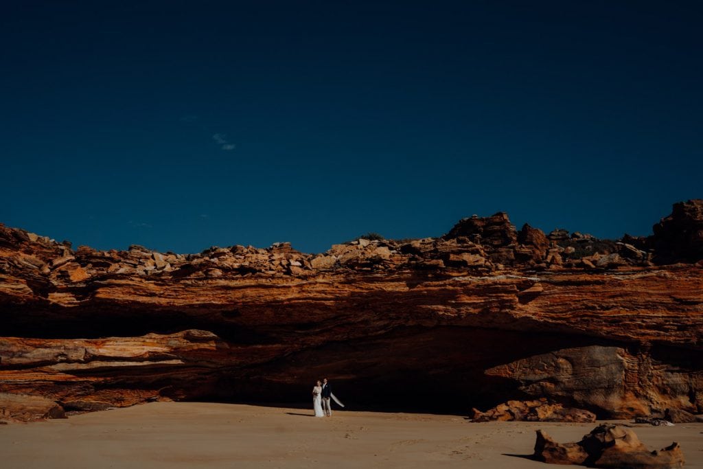 wide shot of wedding couple standing in front of large cave at Eco Beach