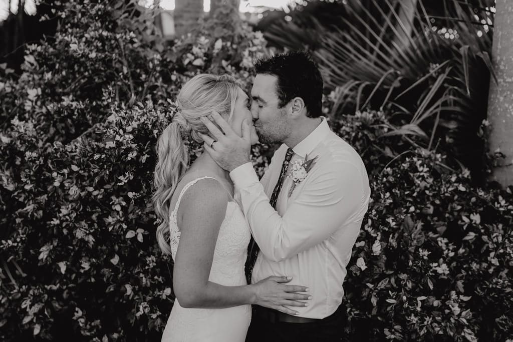 black and white photo of bride and groom kissing in front of a bush with flowers