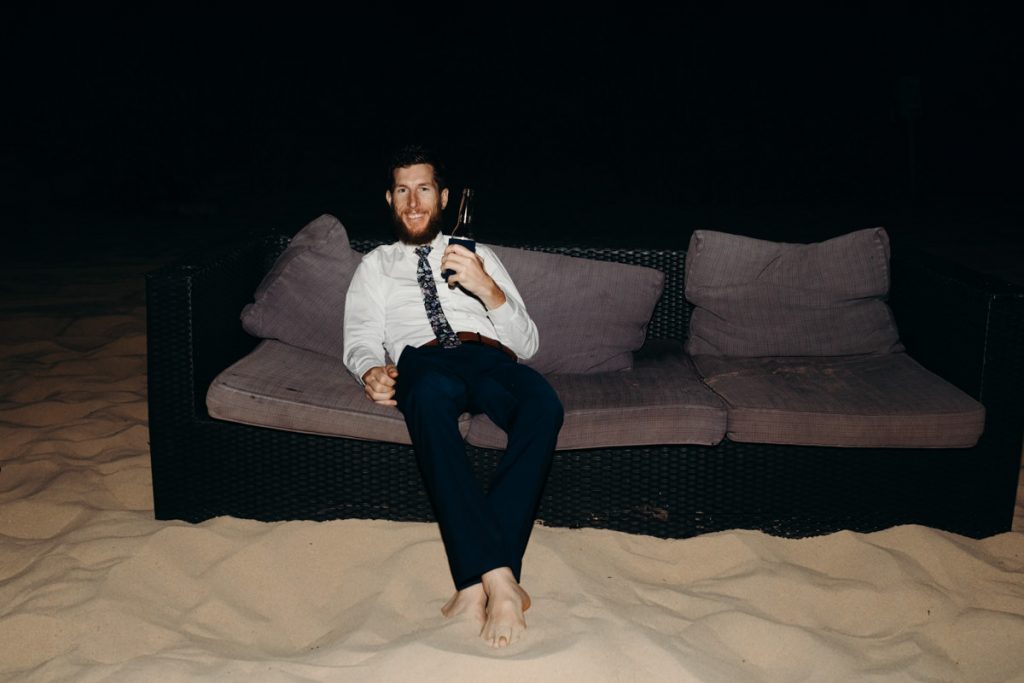wedding guests sits on couch on the beach with beer in his hand