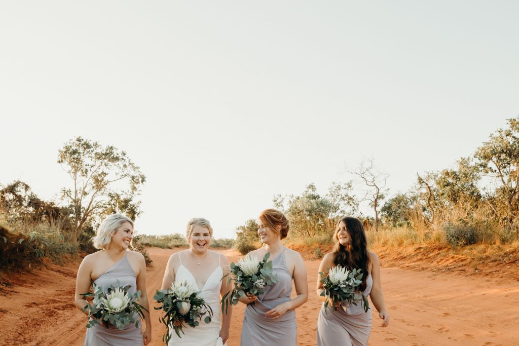 laughing bride with three bridesmaids with flower bouquets walking along on Broome dirt road