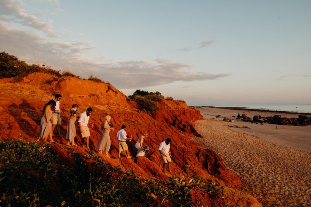 group of people make their way down a red cliff to access Riddell Beach wedding Broome