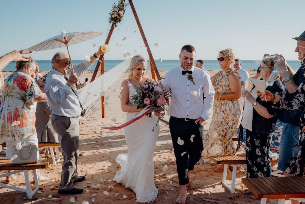 bride and groom walking down the sandy aisle at their relaxed beach wedding with flowers being thrown at them