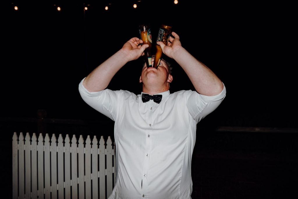 man wearing bow tie and white short drinking two beers at the same time