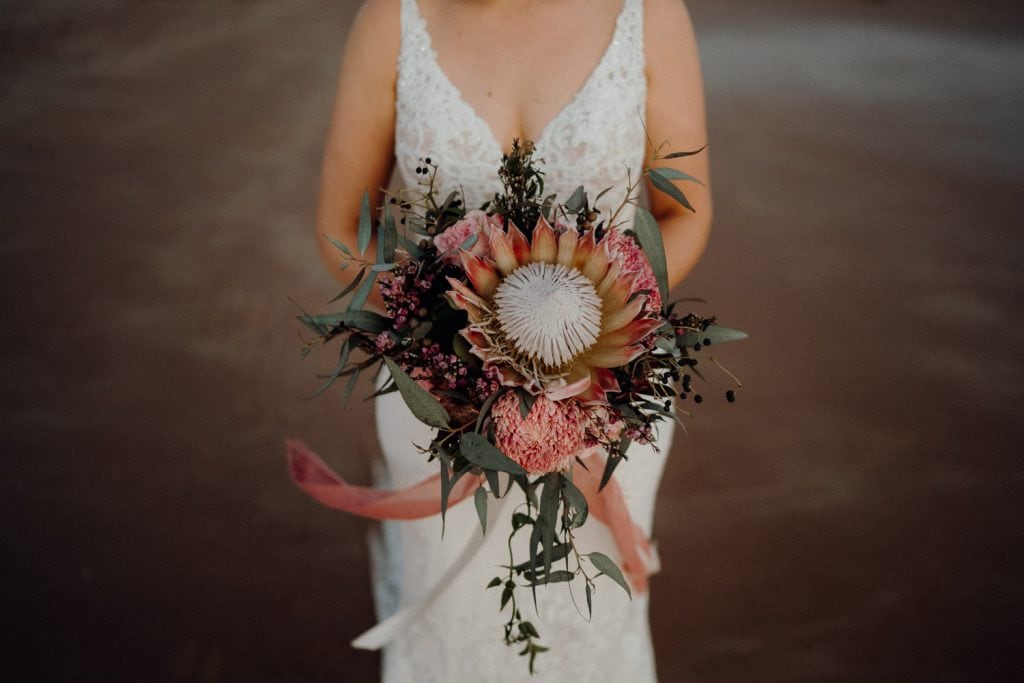 close up of flower bouquet with large protea in the middle held up by bride and white dress