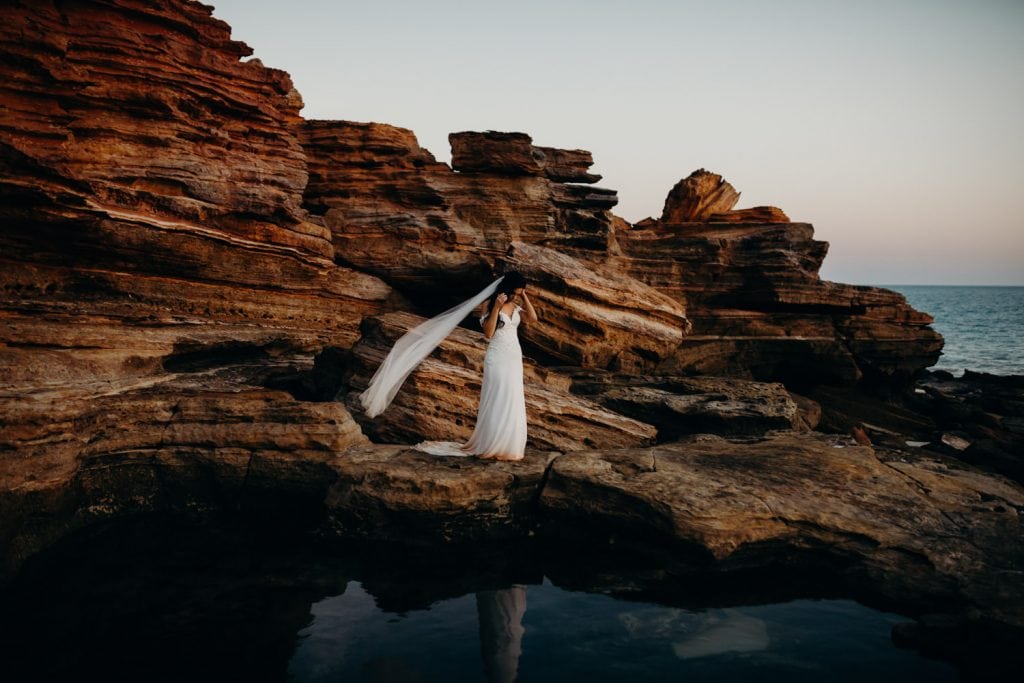 woman wearing white wedding dress stands on rocks with ocean in the background