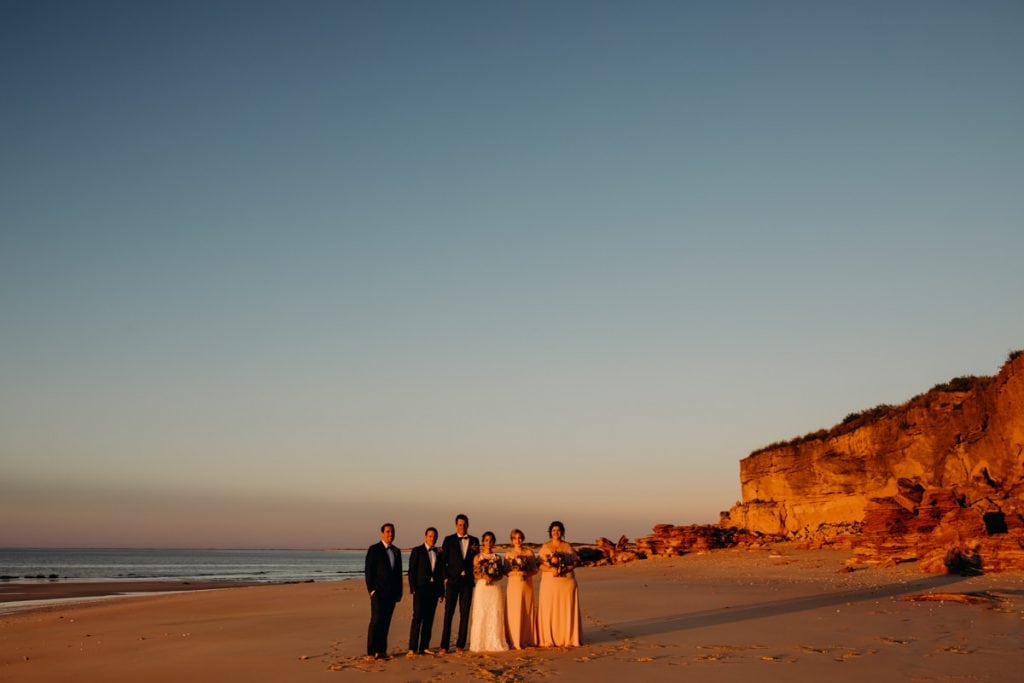 bridal party standing in a line on Eco Beach at sunset with red cliffs in the background